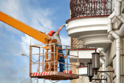 Painter working high in lifting crane with basket repairing historic architecture facade with brush. Worker paints building wall, renovating works. Utility worker use personal protective equipment