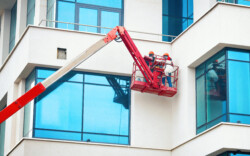 Men at work in hardhat paint facade of the building at height in lifting cradle. Finishing facade work. Construction workers at height working on crane in lifting bucket.